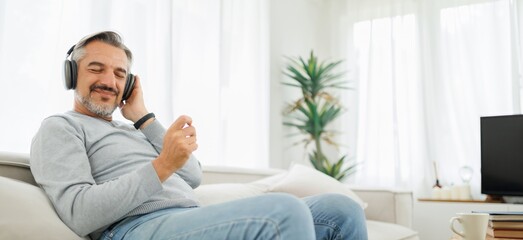 Mature adult man with eyes closed relaxing on sofa while enjoying music through headphones in modern living room.