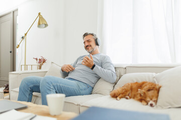 Mature adult man sits comfortably on a sofa and listening to the music with wireless headphones and enjoying with air guitar pose while a pet dog sleeps peacefully nearby.