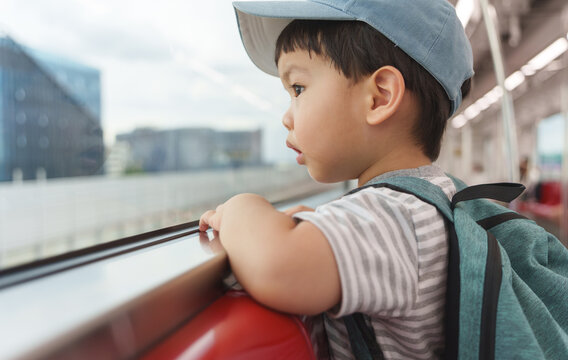 Little Asian boy wearing a blue cap and gray striped t-shirt with backpack standing inside a train while leaning on the window, looking outside with a thoughtful and curious expression.