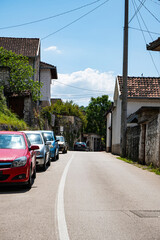 City street features a line of parked cars under a clear blue sky. Traditional stone houses with tiled roofs flank the roadway, creating a serene and picturesque scene.