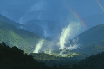 Geysers erupting in a valley between green mountains, forming steam columns under a bright rainbow in a remote location