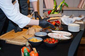 Chefs in white uniforms, black aprons and black gloves stand next to a wooden box with vegetables, tray with herbs, bacon, mushrooms, tomatoes on a black table and are ready to prepare cold appetizers