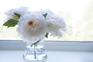 A beautiful bouquet of white artificial peonies stands on a windowsill in a transparent vase and looks like real flowers