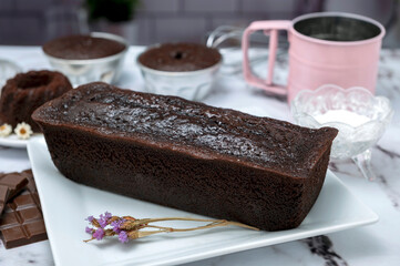 Rectangular and round chocolate pudding with cream on top and strawberries on a table with ingredients and pastry chef's hands decorating