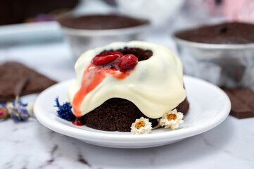 Rectangular and round chocolate pudding with cream on top and strawberries on a table with ingredients and pastry chef's hands decorating