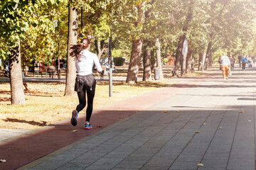 Fototapeta premium A woman jogs along a tree-lined path in a sunlit park. Her motion conveys energy and determination as she navigates the serene environment. Trees and paths create a natural setting.