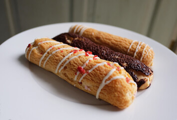 Three long cream puffs, light airy pastries made from choux dough and filled with sweet cream or custard. They have a crispy, golden brown shell, drizzled with white, red, and chocolate icing.