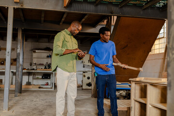 African American coworkers scrolling smartphone and examining plank with tape measure in workshop