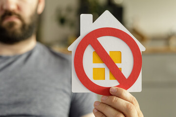 Image of a man holding a house symbol with a red circle and cross, symbolizing homelessness or...