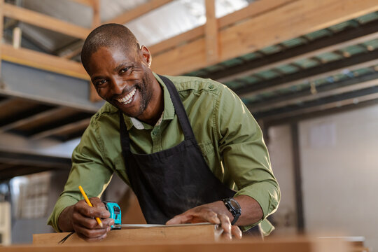 African American man in apron bending over bench marking wood plank with pencil in woodworking shop