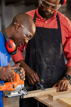 African American male coworkers using jigsaw, holding clamp and cutting plank on workshop bench