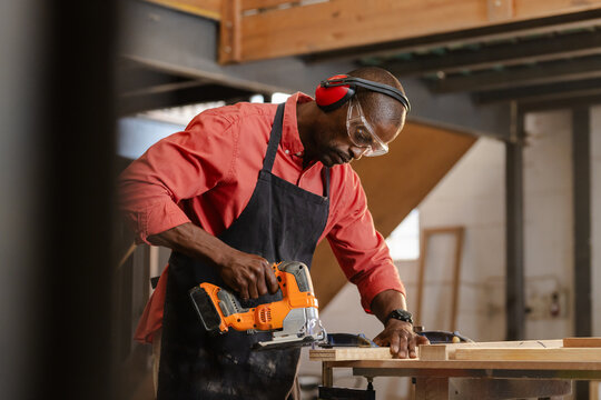 African American man cutting plank at woodshop bench wearing apron, goggles with orange jigsaw saw