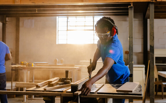 African American male coworkers feeding plank into table saw in carpentry shop wearing earmuffs