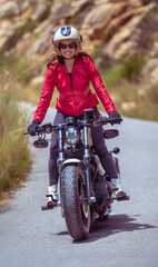 Front view of a smiling young female motorcyclist wearing a red jacket and open-face helmet standing on her custom chopper motorcycle driving along a mountain road on a sunny day.