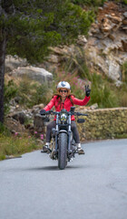 Front view of a young, smiling, and happy female motorcyclist approaching on her classic, customized chopper type motorcycle, making the peace or victory sign with her hand on a mountain road..