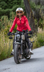 Front view of a smiling and happy young female motorcyclist, wearing a red jacket, riding her customized chopper motorcycle on a steep mountain road, standing on the footrests.