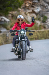 Front view of a smiling and happy young female motorcyclist in a red jacket approaching on her classic customized chopper motorcycle, making the peace or victory sign with her hand on a mountain road.