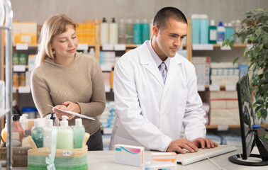 Obraz premium Attentive male pharmacist working in computer while female client standing behind with prescription in drugstore