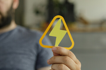A man holds a yellow triangle warning sign depicting a lightning bolt, symbolizing high voltage or electrical hazard.