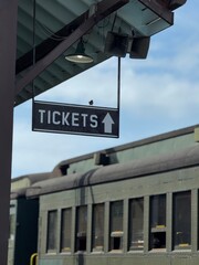 Obraz premium A hanging ticket sign with an arrow, at a train station. A passenger train with windows is visible in the background. A small songbird is perched on the black sign with white letters.