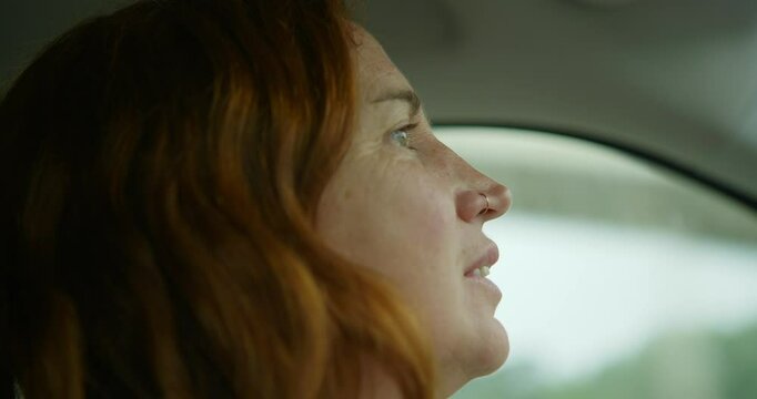 Close-up profile shot of the face of an adult woman with red hair, driving her car, while singing, looking at the road, green eyes, wearing an earring in her nose.  Background green trees.