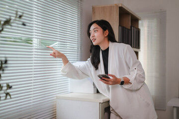 Businesswoman opening blinds and holding smartphone in office