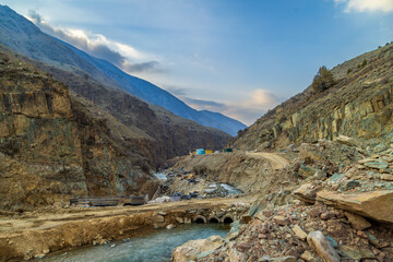 Serene mountainous region features a river flowing through rugged terrain. Hydroelectric node construction site with pipes and small buildings indicates ongoing development in this desolate outdoor