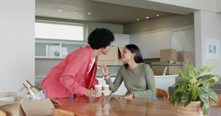 Diverse couple sitting at dining table among moving boxes, exploring new home while sharing coffee - Powered by Adobe