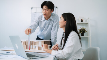 Professional male asian architect using ruler to measure house model length while young beautiful caucasian colleague using laptop to analyzed data on meeting table with house model. Immaculate.