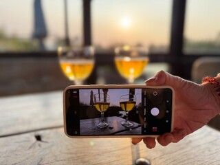 A smartphone captures two glasses of drink on a table during sunset. Creative perspective highlights technology, leisure, photography, and the beauty of shared moments in warm evening light.