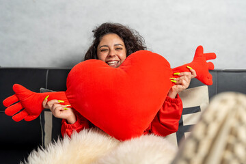Young woman holding hugging heart shaped pillow smiling at home