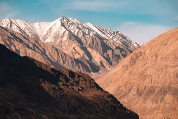 Fototapeta premium Golden sunlight casts long shadows across the dramatic Nubra Valley landscape, revealing rugged mountain textures and snow-capped Himalayan peaks under a vast blue sky in northern Ladakh, India
