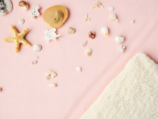 A soft flat lay of various seashells and coral fragments placed on a textured pink towel, next to a cream-colored mesh beach bag.
