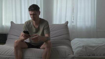 Young man reacts with disgust to something on his phone while sitting on a sofa in a modern apartment. - Powered by Adobe