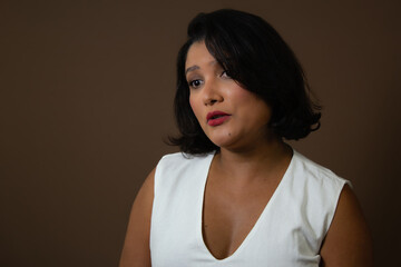 Worried brown skin woman wearing a white tank top, talking to someone. Ideal for concern, worries, discussion. Horizontal studio portrait with copy space on brown background.