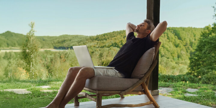 A man sits outdoors with a laptop on his lap, enjoying a relaxed moment while working remotely in a natural setting. Work-life balance concept. Space for text