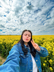 Carefree female tourist taking smartphone selfie in spectacular yellow flower field. Rapeseed flowers. Great for social media aesthetics, positivity campaigns, and springtime visuals.