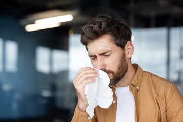 A man with a beard blowing his nose with a tissue, possibly suffering from allergies or a cold.