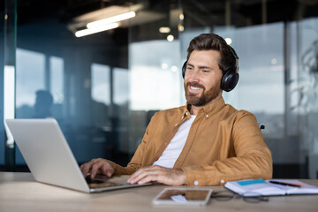 A man with a beard wearing headphones is smiling while working on a laptop at a wooden desk.