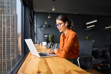 Busy latin hispanic young business woman working on laptop computer sitting at office desk with city view. Indian entrepreneur manager businesswoman using pc for communication, learning at workplace