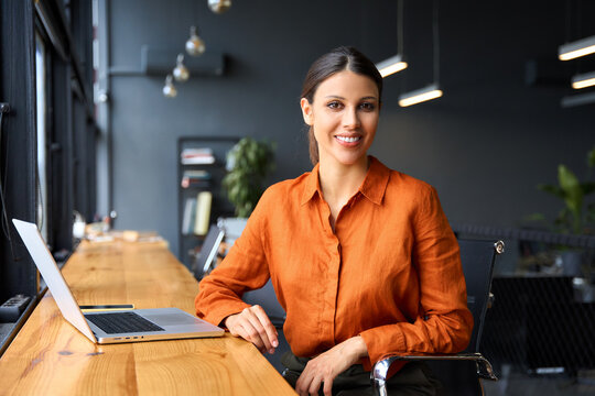 Portrait young it specialist latin hispanic business lady working on laptop pc sitting at desk in modern office smiling at camera. Middle eastern indian woman using computer technology for work online