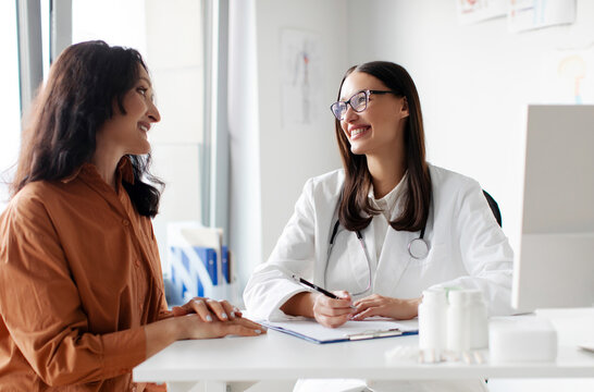 Friendly woman doctor therapist and female patient sitting and talking, looking at each other in medical clinic office during consultation