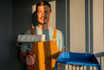Young painter stands confidently in a sunlit room, holding a paint roller and tray. Dressed in overalls, he is ready to start a home renovation project, embodying creativity and craftsmanship © wifesun