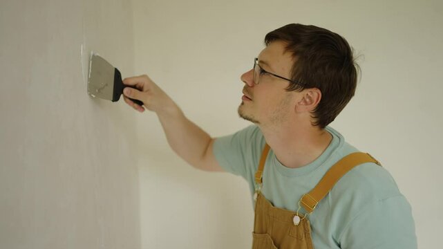 Professional house painter wearing glasses and brown overalls, filling cracks in a white wall using a putty knife and plaster before starting to paint