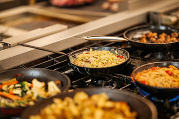 Variety of hot dishes including pasta, vegetables and roasted potatoes cooking in skillets on a gas stove. Close-up of active food preparation in a commercial kitchen.