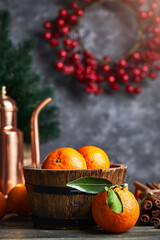 Still life with copper pitcher teapot and ripe fresh orange tangerine fruits in a wooden, barrel-shaped rustic bowl on bark wood table. Grunge stone wall berries wreath Christmas tree on background.