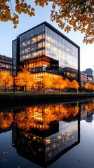 A modern, glass-fronted office building, illuminated internally, is reflected in a calm canal at twilight. Autumnal trees with golden leaves frame the scene
