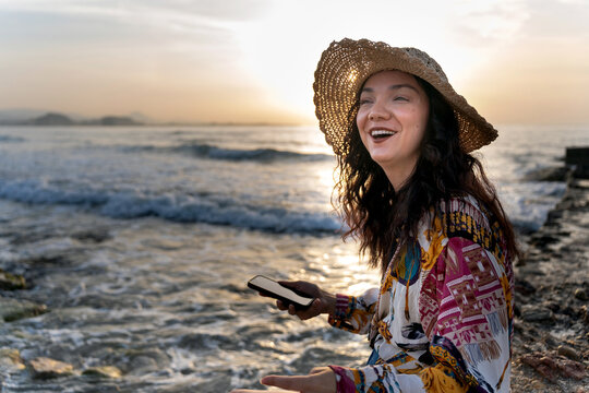 Romanian woman enjoying sunrise at the beach with smartphone and straw hat