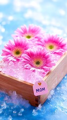 Three pink gerbera daisies nestled in a light wood box filled with ice, a small heart-shaped tag reading "MOM" adorns the box, set against a light blue background