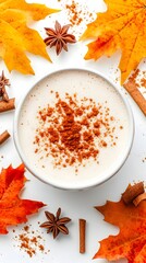 Overhead view of a creamy beige beverage in a white bowl, topped with cinnamon powder.  Surrounding it are autumnal orange and yellow maple leaves, cinnamon sticks, and star anise. The scene is set